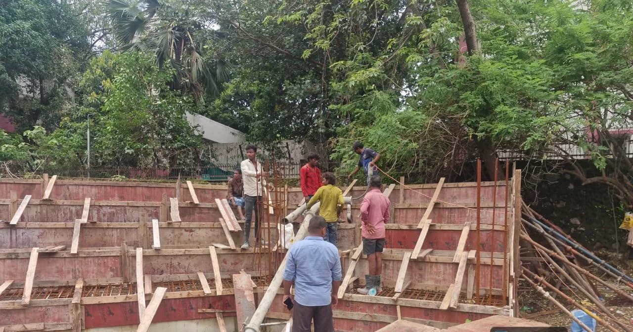 A crew setting timber formwork against a retaining wall at the Kharghar park site, with a concrete line running in