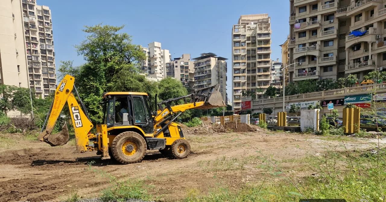 A backhoe grading the cleared park site at Kharghar, residential towers along the far edge