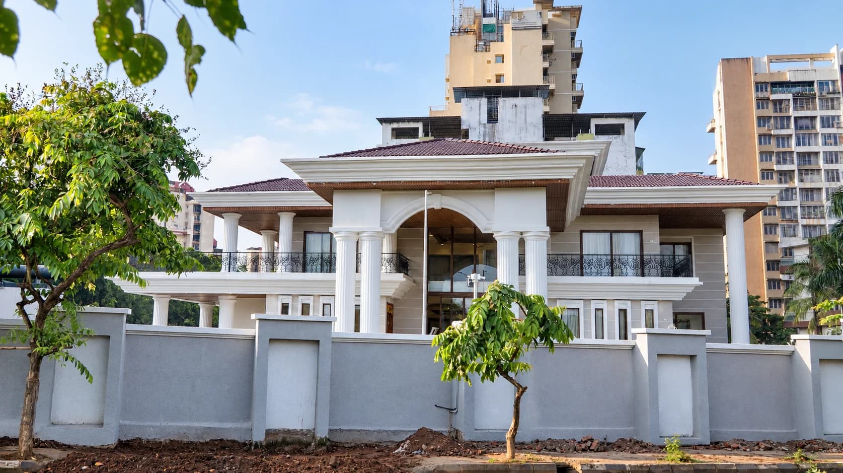 A white colonnaded two-storey villa with a tiled roof behind a boundary wall, young trees in front and apartment towers beyond