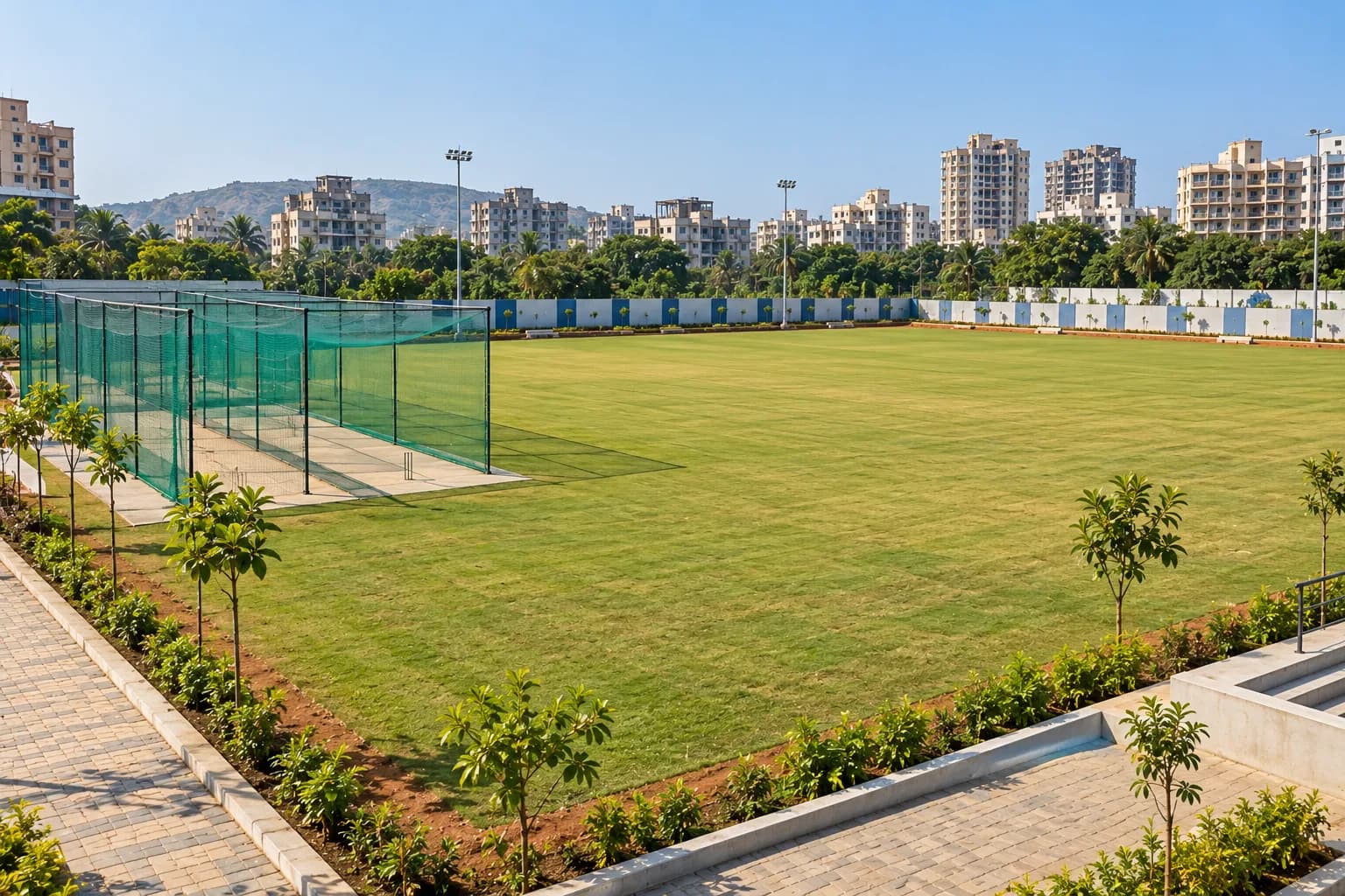 A developed municipal playground: practice cricket nets to one side, open mown lawn across the centre, a painted compound wall along the boundary, housing beyond