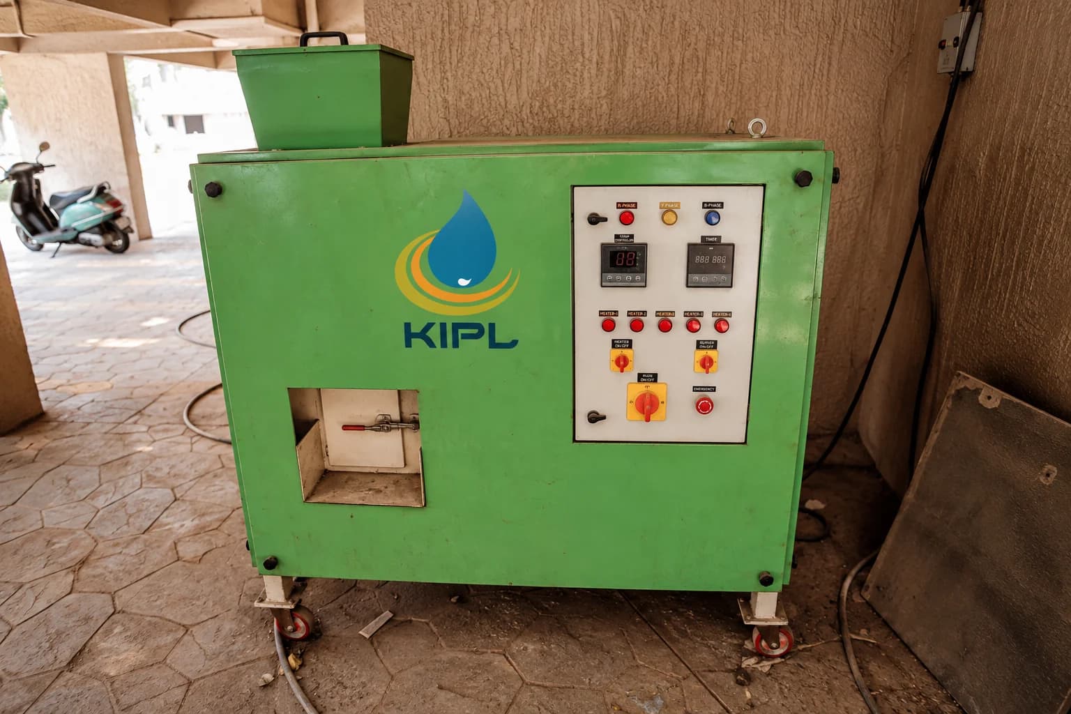 A green KIPL organic waste converter on castors under a building podium, feed hopper above and control panel to one side